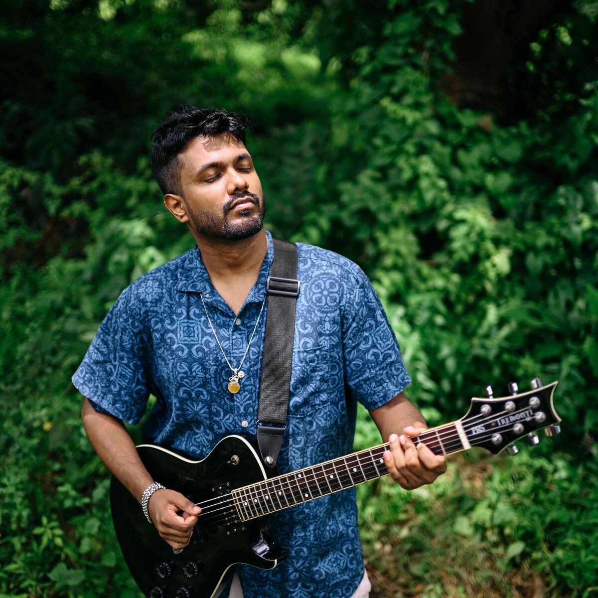 Ashwin Syam playing an electric guitar among green leaves, eyes closed